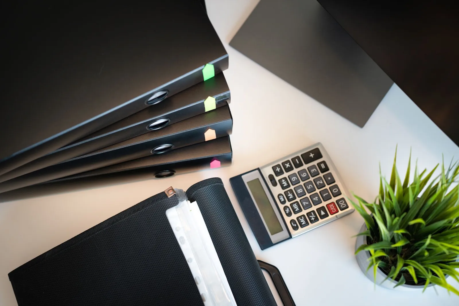 Black folders with coloured tabs, a calculator and a pot plant on a white desk, photographed from above