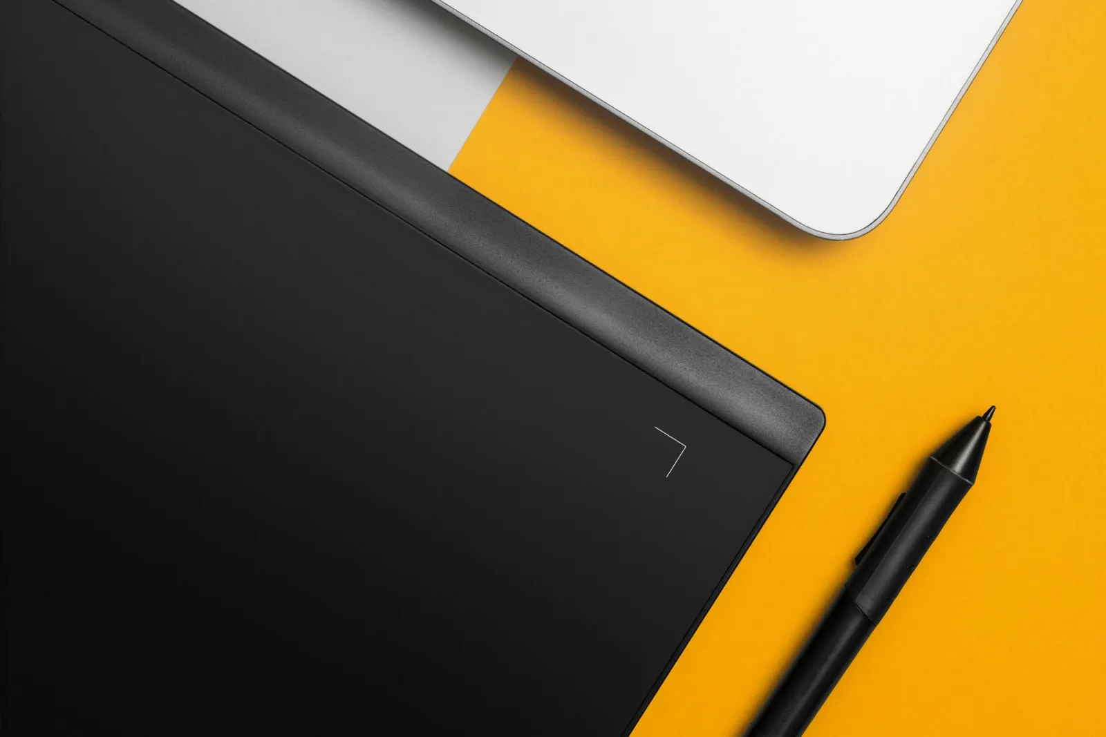 Black drawing tablet with stylus and part of a laptop on a yellow background, photographed from above